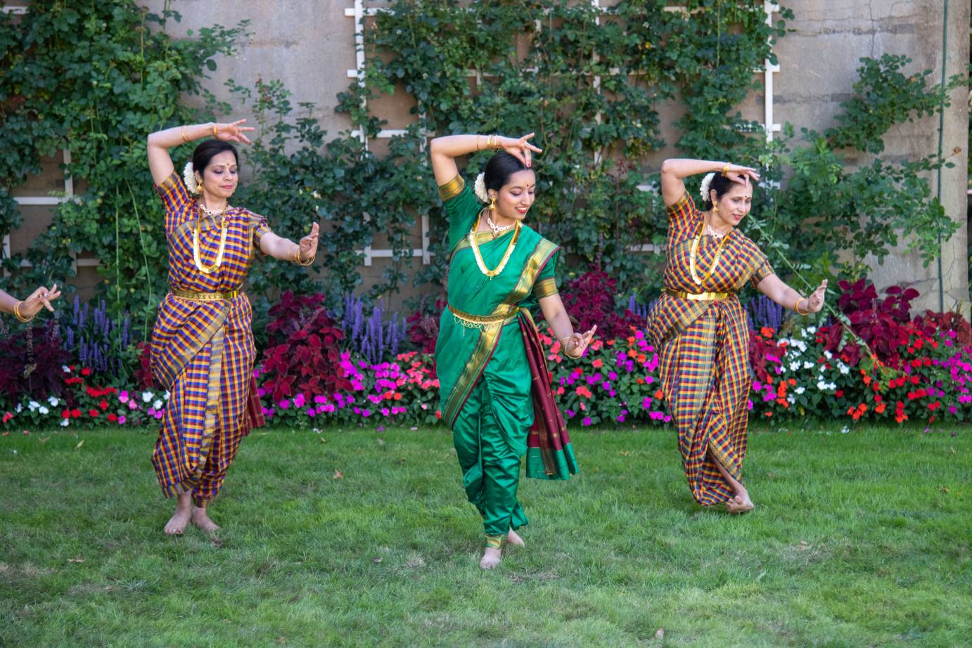 Three Kathak dancers in brightly colored traditional dress with gold belts and necklaces enliven the lawn background with more color. the dancer in the foreground wears bright green and gold, the dancers in the background wear checked prints
